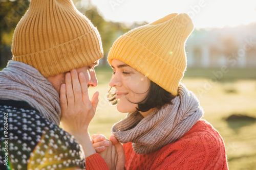 Horizontal portrait of brunette female in warm hat, scarf and sweater, keeps hands on boyfriend`s cheek, have romantic moment, going to kiss as dating. Couple in love look at each other passionately photo