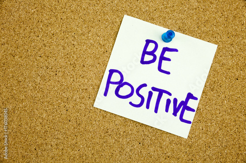 A yellow sticky note writing, caption, inscription be positive, motivational lifestyle reminders against black ext on a colorful sticky note pinned to a cork notice board photo