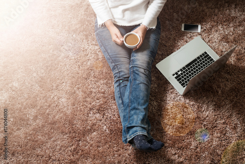 Woman holding cup of coffee sitting on carpet online with laptop and smartphone,aerial view. Coffee relaxation in living room.
