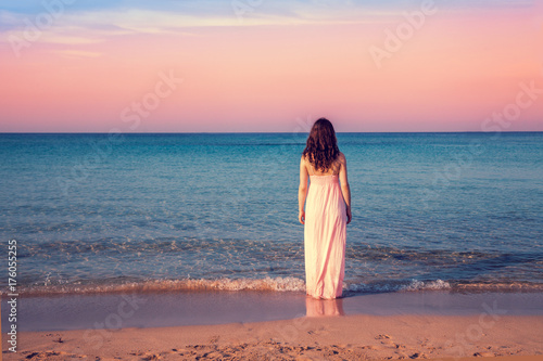 Young woman in a long dress back to camera at sunset on the beach