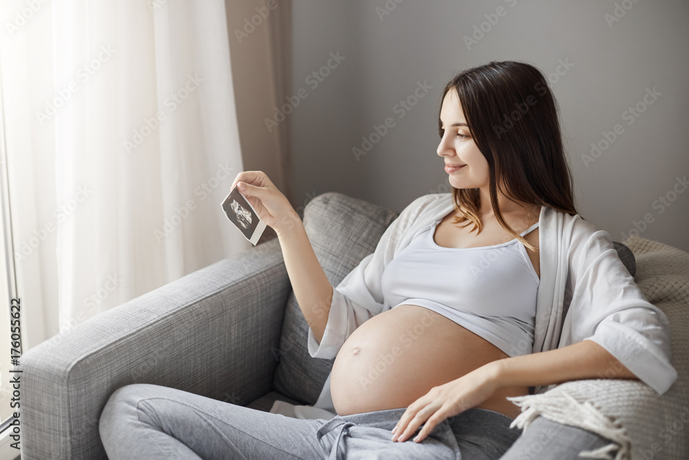 Pregnant lady looking at a baby ultrasound picture trying to guess is it a boy or a girl. Staying home in her cozy sofa.