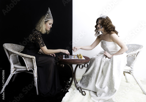 Two girls wearing queen dress; diamond crown and other jewelry sitting in chairs opposite to each other and playing chess. 
Competition; opposition concepts. Black and white background photo