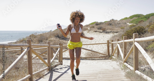 Young ethnic woman in summer sportive clothing holding smartphone with headphones listening to music while dancing on pier on beach. © Dash