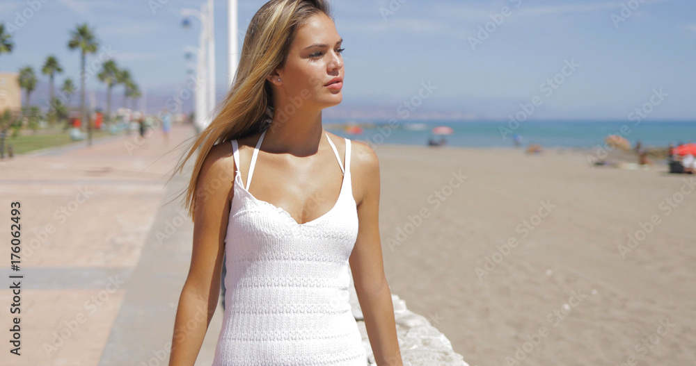 Young blodne in white beach garment enjoying stroll alongside tropical beach while posing at camera in bright sunlight. Young blodne in white beach garment enjoying stroll alongside tropical beach while posing at camera in bright sunlight.