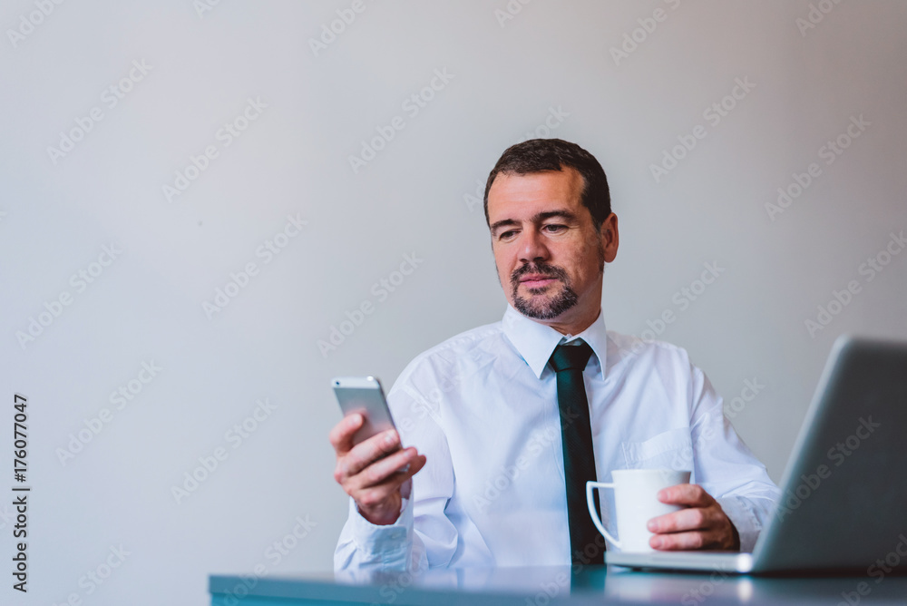Mature businessman in suit using cell phone at desk in small office. Mature businessman in suit using cell phone at desk in small office.