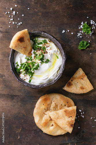 labneh middle eastern lebanese cream cheese dip with olive oil, salt, herbs served with traditional pita bread in terracotta bowl over dark texture wooden background. Top view with space