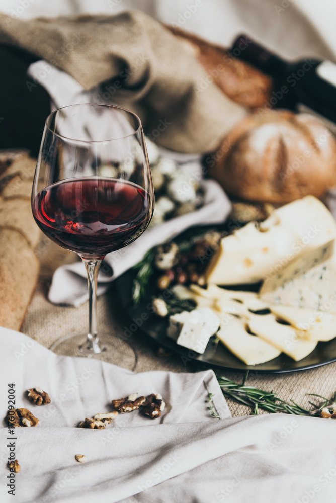 A wineglass is filled with dry red wine lying behind. Fresh bread, blue cheese, masdaam cheese, quail eggs and nuts are used as decoration.