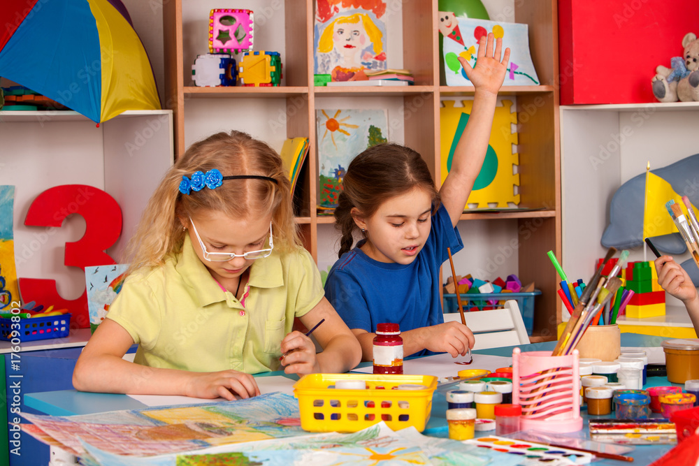 Fototapeta premium Small students painting in art school class. Schoolgirl raises her hand to answer.