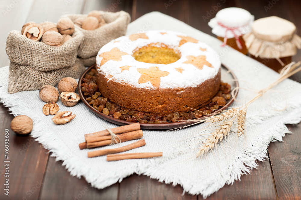 homemade traditional fruit cake on clay plate decorated with wheat ears, nuts, cinnamon and honey jars at wooden table