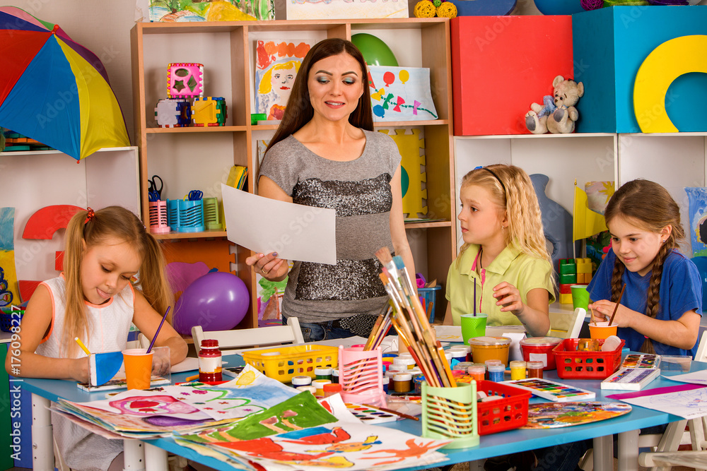 Fototapeta premium Kids playroom organization of children painting and drawing in kid's club. They learn to live in a team. Kindergarten teacher and students work together. Teacher praises charges for good drawings.
