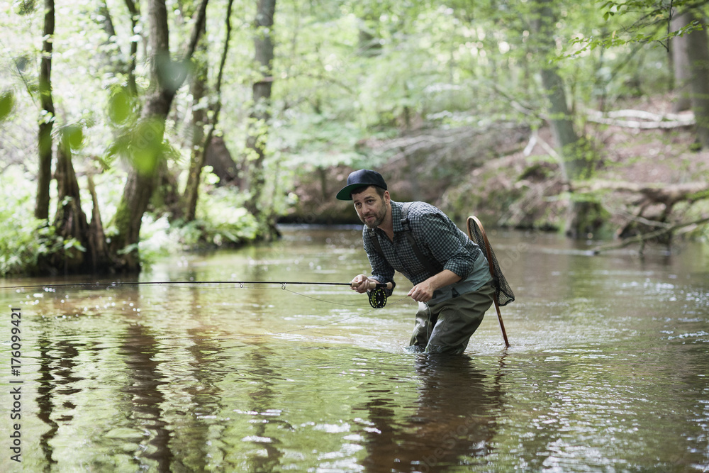 A patient man in waders is fly fishing on a river in forest area. A patient man in waders is fly fishing on a river in forest area.