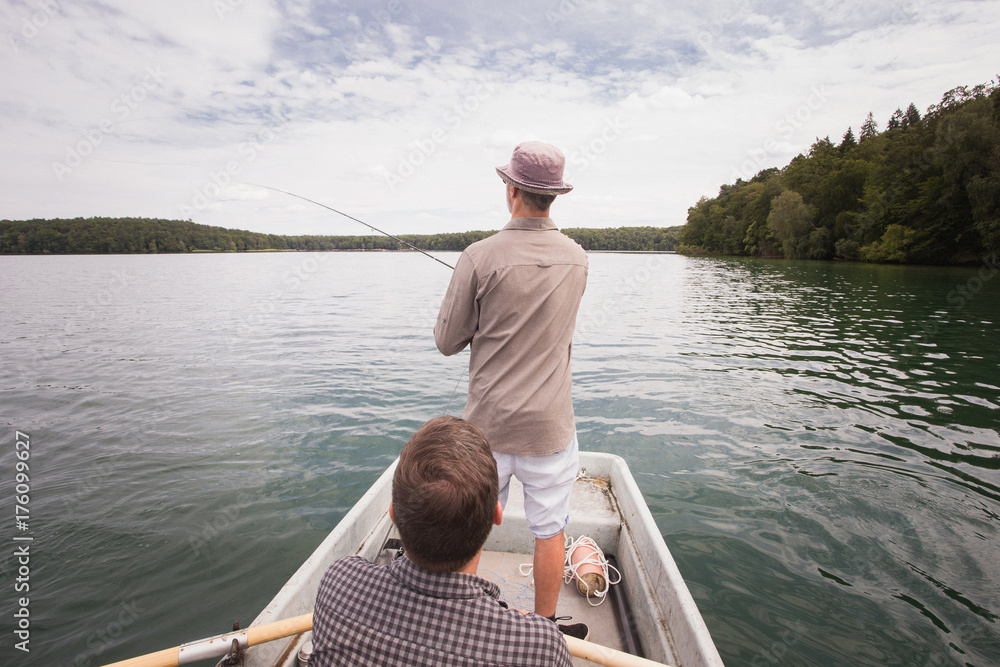 A man is rowing a rowboat while his friend is fly fishing from the boat on a lake. A man is rowing a rowboat while his friend is fly fishing from the boat on a lake.