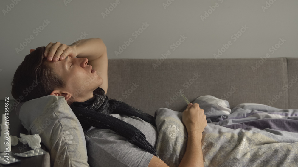 Ill young man is taking temperature and coughing in his bed. Plenty pills and medications are on the bedside table Ill young man is taking temperature and coughing in his bed. Plenty pills and medications are on the bedside table
