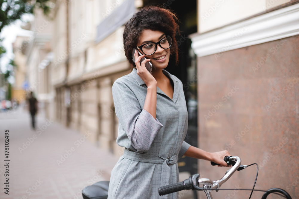 Portrait of cheerful African American girl in glasses standing with bicycle and talking on her cellphone. Young beautiful lady with dark curly hair in gray dress standing with bicycle on street