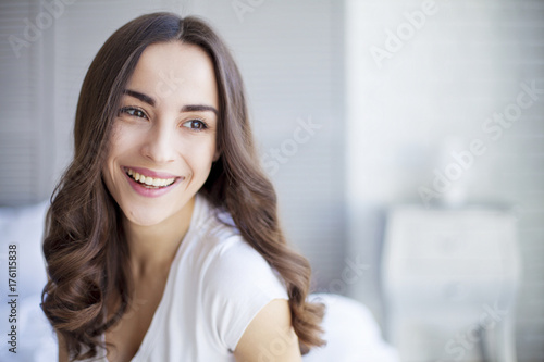 Portrait of Beautiful young brunette woman sitting at the end of the white bed and smiling.