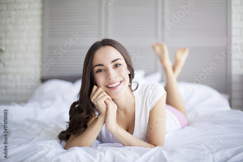 Attractive young smiling brunette woman lying in white bed and looking at camera in her bedroom.