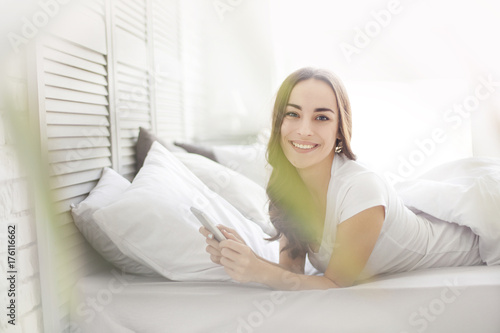 Smiling side portrait of beatiful female laying relaxing on home bed holding smart phone with blank screen, networking, indoors bedroom. Brunette woman using technology, lifestyle, home interior.