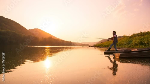 A cute teen catches a fish from a wooden boat on the river. The sun is hiding behind the mountain