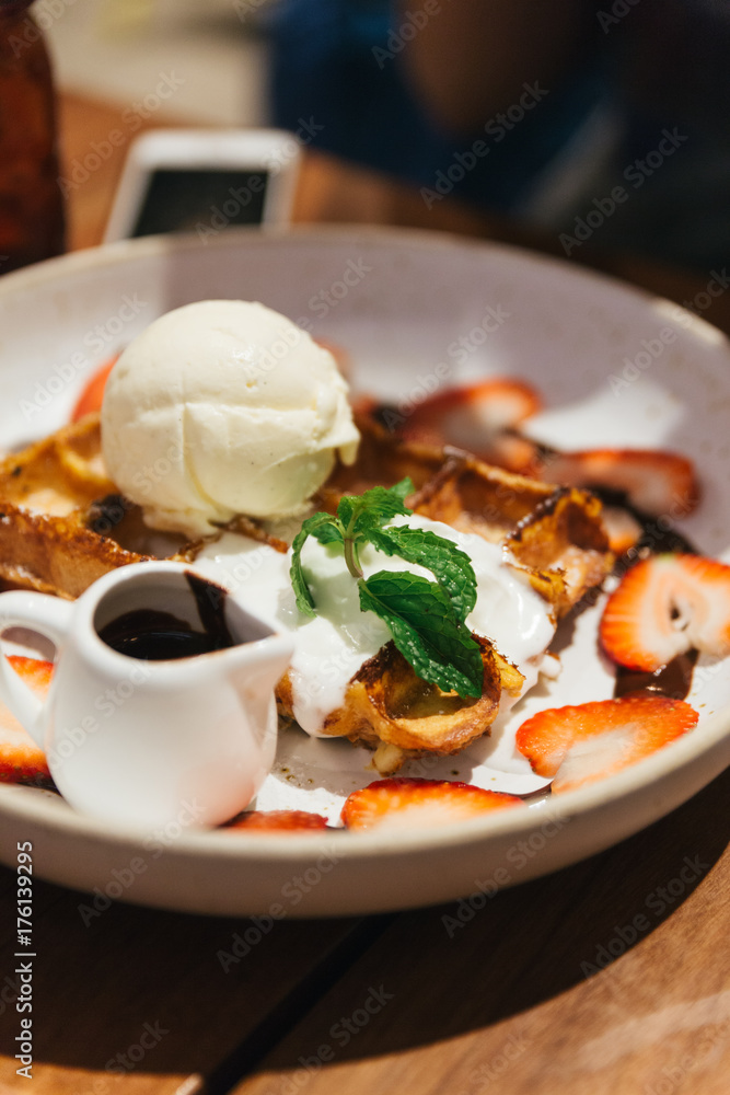 Waffle topping with a scoop of vanilla ice cream, whip cream, sliced strawberry, mint leaves and chocolate sauce on white plate at dessert cafe in Bangkok, Thailand. Waffle topping with a scoop of vanilla ice cream, whip cream, sliced strawberry, mint leaves and chocolate sauce on white plate at dessert cafe in Bangkok, Thailand.