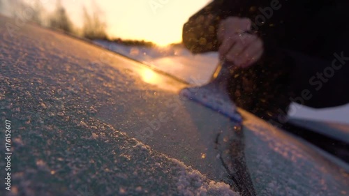 SLOW MOTION CLOSE UP DOF: Person cleaning morning frost off a car window at golden sunrise. Hand scraping hoarfrost off a frozen car windshield, snowflakes flowing over the sun. Scraper removing frost