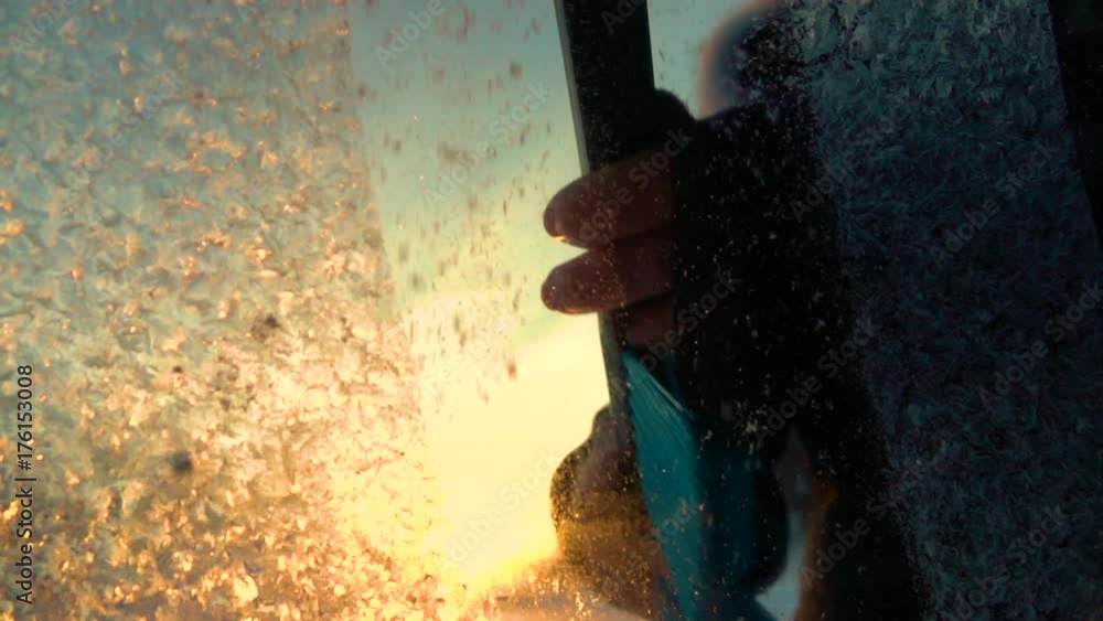 SLOW MOTION CLOSE UP DOF: Person cleaning morning frost off a car window at golden sunrise. Hand scraping hoarfrost off a frozen car windshield, snowflakes flowing over the sun. Scraper removing frost