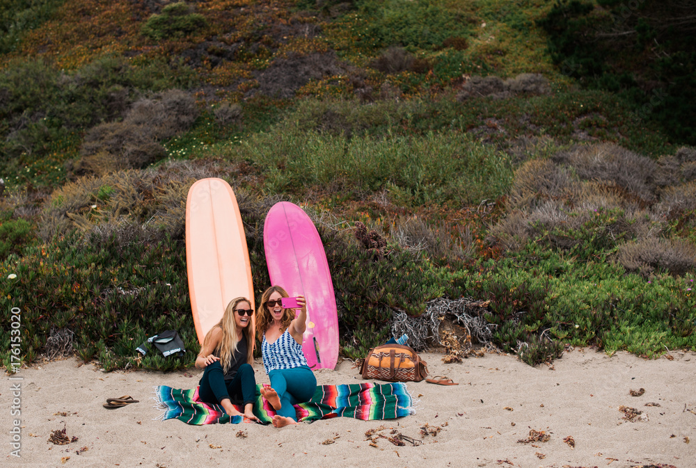 Two Young Woman Sitting on the Beach Taking Selfie Photographs Two Young Woman Sitting on the Beach Taking Selfie Photographs
