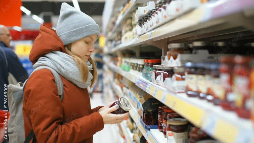 Young Lady is Comparing to Jars with Jam in a Supermarket Young Lady is Comparing to Jars with Jam in a Supermarket