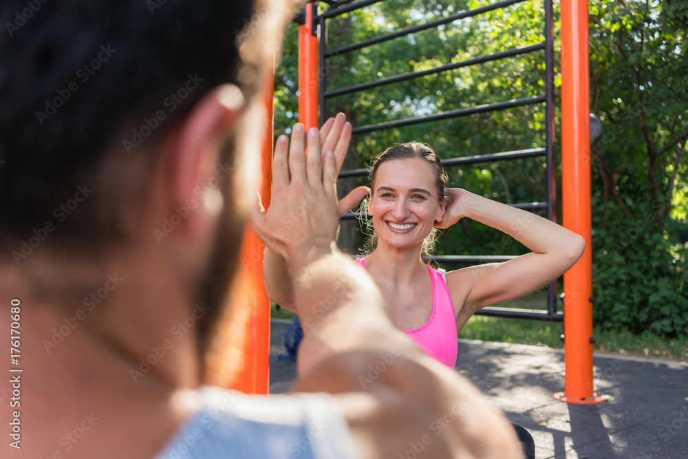Obraz premium Over the shoulder view of a cheerful young woman giving high-five to her partner while doing crunches outdoors, during couple workout in a fitness park
