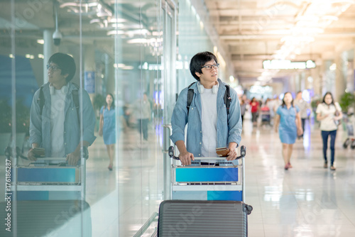 Young Asian man walking with airport trolley and his suitcase luggage in the international airport terminal, arrival from travel abroad