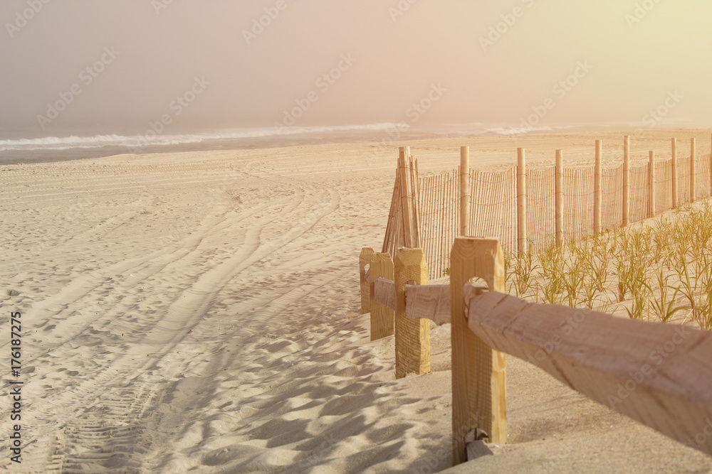 Empty beach at lonely oceanside with warm emotional bright sun pouring into the frame.  Wooden railing and waves in the distance.  Copyspace room for text.
