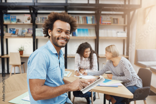 Close up portrait of good-looking black-skinned university student sitting on meeting with friends after study, talking about group project, looking through websites on digital tablet.