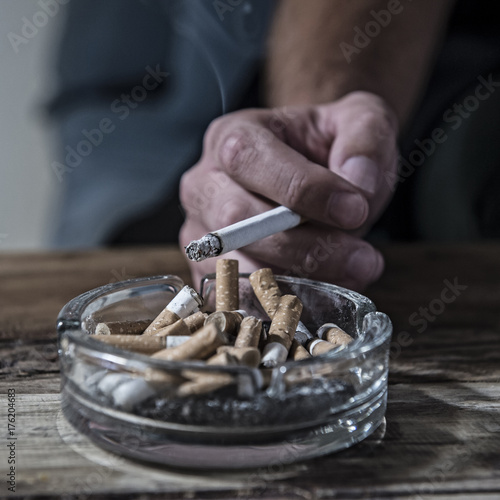 hand of man smoking cigarette with ashtray full of cig butt end and ash in disgusting unhealthy addiction