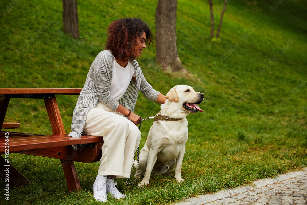 Young african lady sitting on bench in park and holding dog Young african lady sitting on bench in park and holding dog