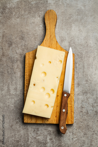 Tabla con queso emental en trozo sobre una tabla de madera rústica y un cuchillo sobre un fondo gris texturado. Vista superior y de cerca. Formato vertical