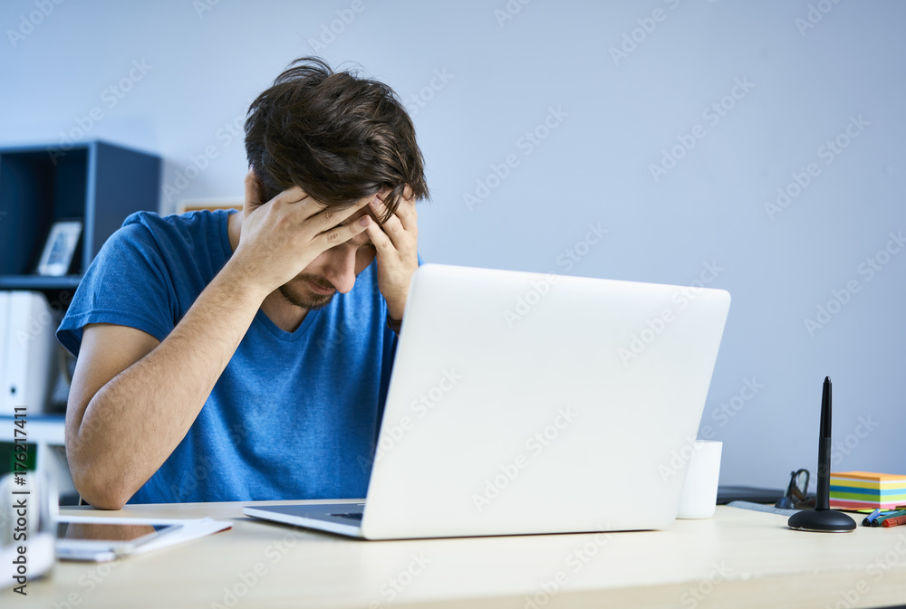 Tired young man in front of laptop in home office having headache Tired young man in front of laptop in home office having headache