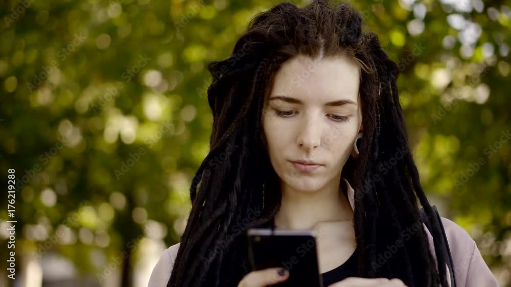 teenage girl with informal hairstyle is standing in a park and looking on a screen of phone teenage girl with informal hairstyle is standing in a park and looking on a screen of phone