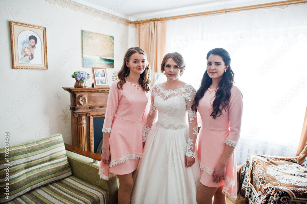 Fabulous young bride posing with two gorgeous bridesmaids in pink dresses in light room.
