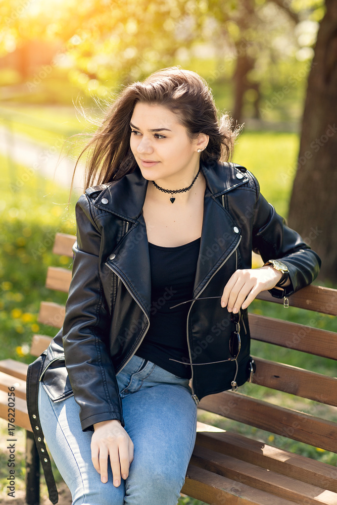 Glamorous young Caucasian woman in black leather jacket sitting in the park on the bench Glamorous young Caucasian woman in black leather jacket sitting in the park on the bench