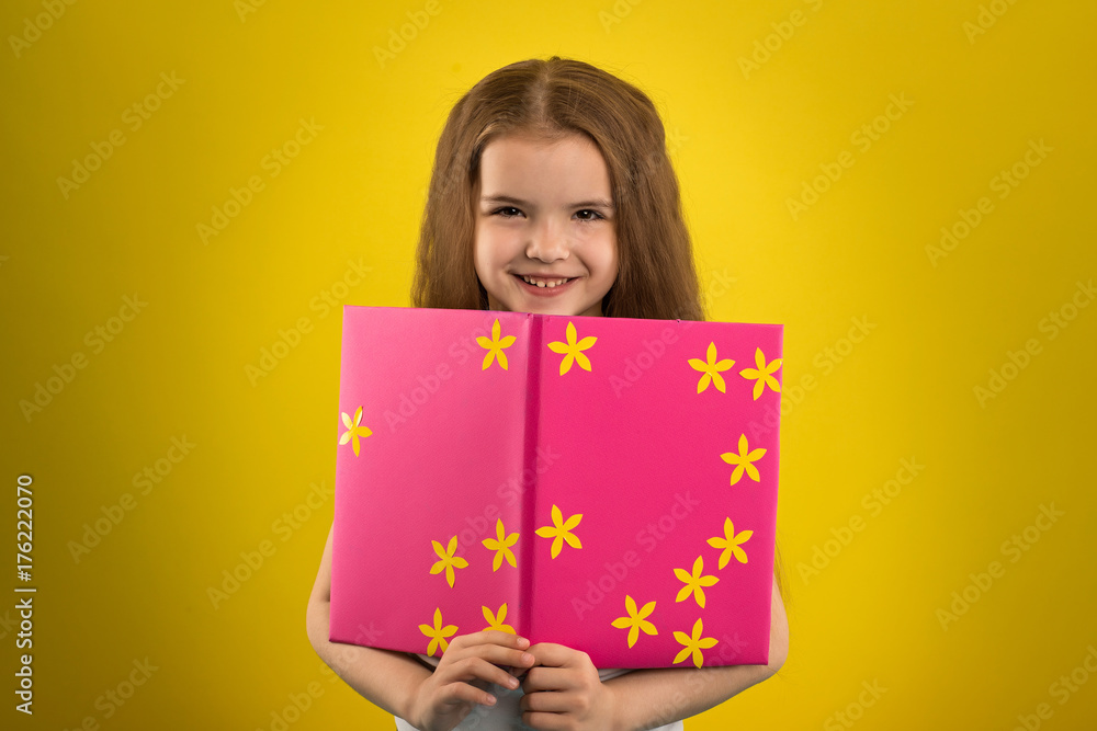 Concept - education. Girl reading a book in a pink cover, shooting in the studio on an isolated yellow background Concept - education. Girl reading a book in a pink cover, shooting in the studio on an isolated yellow background