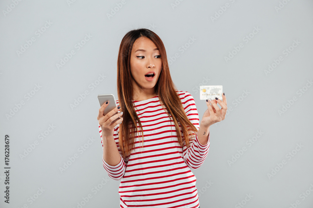 Shocked asian woman in sweater using smartphone and credit card Shocked asian woman in sweater using smartphone and credit card