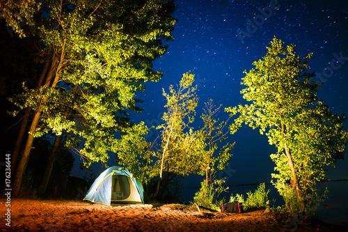 Tent set on the sandy beach among the trees on coast of a river with stars in the night sky