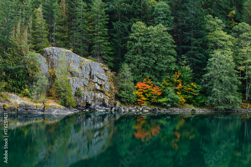 Autumn colors by a peaceful lake with a large rock formation and forest background.