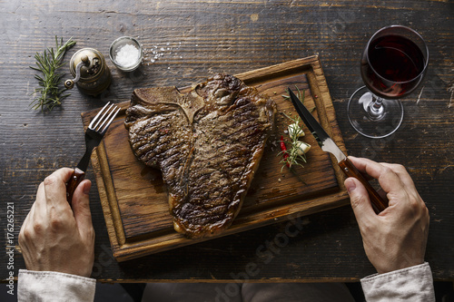 Grilled T-bone steak on serving board, fork and knife in male hands and red wine on wooden table background photo