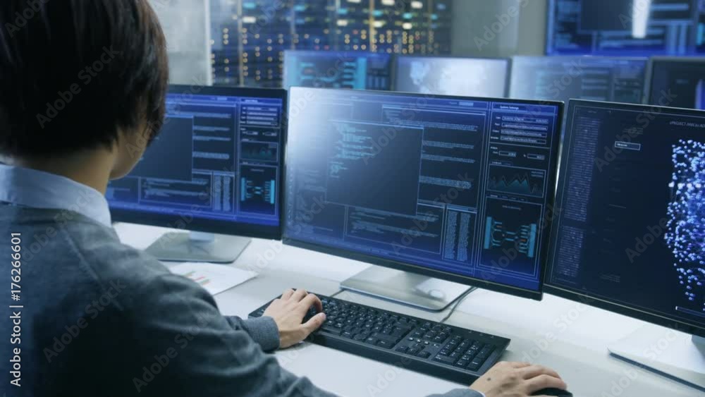 In the System Control Room Technical Operator Works at His Workstation with Multiple Displays Showing Graphics. Possible Power Plant/ Airport Dispatcher/ Data Center/ Government Surveillance/ Space. In the System Control Room Technical Operator Works at His Workstation with Multiple Displays Showing Graphics. Possible Power Plant/ Airport Dispatcher/ Data Center/ Government Surveillance/ Space.