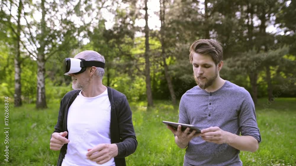 Man with tablet and senior father with VR glasses outdoors. Man with tablet and senior father with VR glasses outdoors.