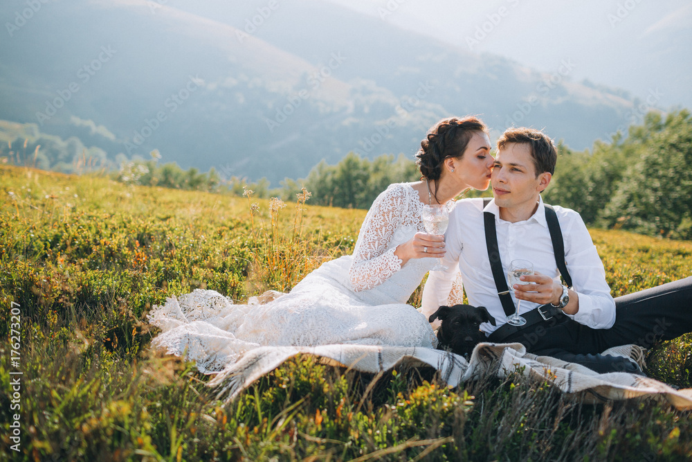 Bride and groom drink champagne sitting with puppy on the hill Bride and groom drink champagne sitting with puppy on the hill