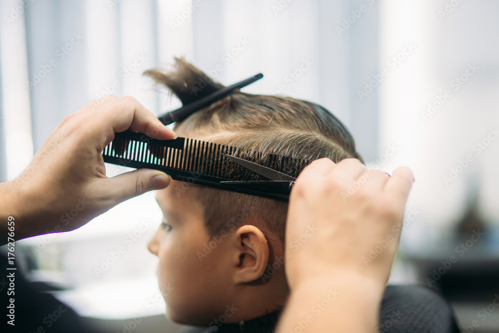 Fototapeta premium Little Boy Getting Haircut By Barber While Sitting In Chair At Barbershop.
