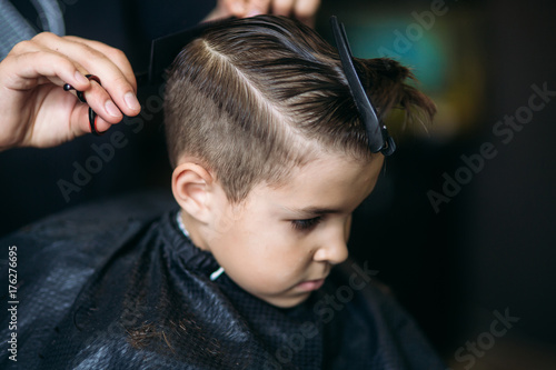 Fototapeta Mały chłopiec uzyskiwanie fryzury przez fryzjer, siedząc w fotelu w fryzjera.