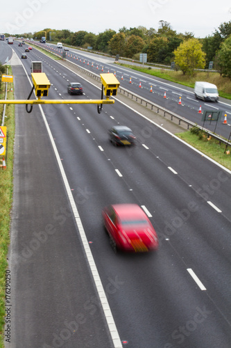 A pair of Average Speed Check cameras above a busy motorway or highway in the UK with speeding traffic below including cars, vans and trucks.