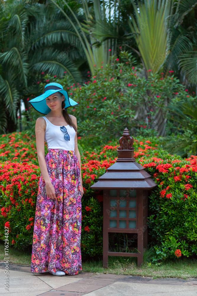 A girl in azure hat with fields, a white tshirt and a long pink skirt, against the background of a Chinese lantern and a flower bed. Sanya, Hainan Island, China. A woman in Sanya, Hainan, China.